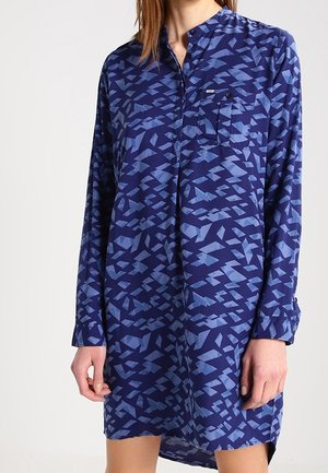 Vrouw draagt een blauw overhemdjurk met lange mouwen, met een geometrisch patroon en een borstzakje met knoop.