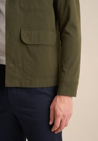 Olivgrüne leichte Jacke mit zwei vorderen Taschen, geradem Schnitt und langen Ärmeln, kombiniert mit navyblauen Hosen.