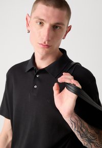 Juodas trumpas rankovės polo marškinėliai su trimis mygtukais ir išsiuvinėtu logotipu. Modelis laiko juodą diržą su tekstūruotais akcentais, demonstruodamas kūno meną.