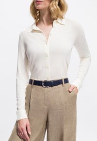 Donna che indossa una camicia a maniche lunghe di colore crema, con bottoni, infilata in pantaloni beige a vita alta con una cintura nera, con la mano in tasca.