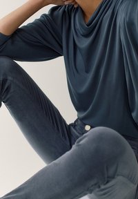 En person sitter med en arm vilande på det böjda knät, iklädd en lös blå långärmad topp och åtsittande blå jeans mot en enfärgad bakgrund.