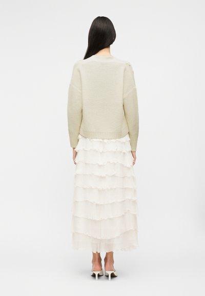 Γυναίκα με μακριά σκούρα μαλλιά, ντυμένη με εκρού πλεκτό πουλόβερ, λευκή φούστα με βολάν σε στρώσεις και λευκά mule με τακούνι, στεκόμενη με την πλάτη προς τα εμάς σε απλό φόντο.