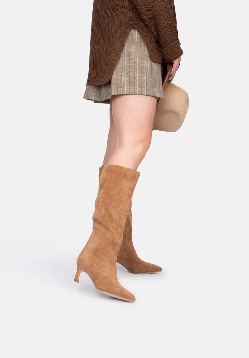 Botas de ante en color tostado hasta la rodilla, con punta afilada y tacón delgado. Acompañadas de una falda de cuadros, que muestra una apariencia texturizada y suave.