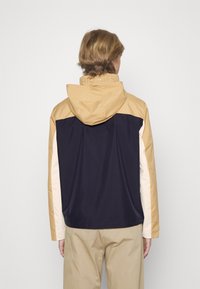 Farveblokket jakke med hætte, der har paneler i beige, marineblå og creme. Glat, letvægtsstof med et moderne design.