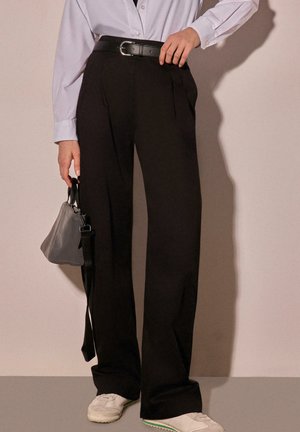 Person wearing black wide-leg trousers with a black belt, white sneakers, white button-up shirt, holding a small gray handbag by their side.