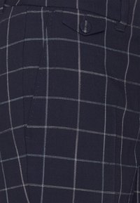 Mörkblå mönstrad tyg med rutmönster, här med ljusblå och vita linjer, strukturerad yta, detaljerad ficka med knapp.