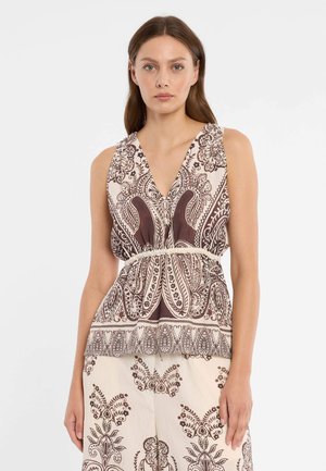 Vrouw draagt een mouwloze top met V-hals en bijpassende broek met ingewikkelde crème- en donkerbruine bloemmotieven, staande tegen een effen achtergrond.