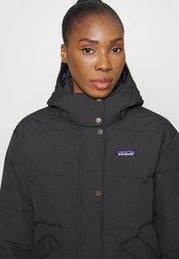 Black puffer jacket featuring a hood, snap buttons, and a Patagonia logo patch. The material is textured with horizontal quilted sections.