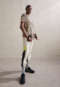 Top s krátkým rukávem s leopardím potiskem, krémové tepláky s černými a neonově zelenými akcenty, černé tenisky, šedé ponožky, texturované pozadí.