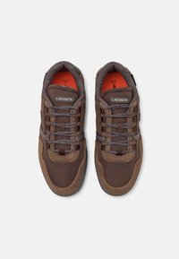Braune Sportschuhe, die eine Mischung aus strukturiertem Wildleder und Netzmaterial aufweisen, mit grauen gemusterten Schnürsenkeln und leuchtend orangefarbenem Innenfutter.