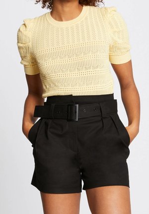 Femme portant un haut en maille jaune à manches courtes avec des épaules bouffantes et un short noir ceinturé taille haute, les mains dans les poches.