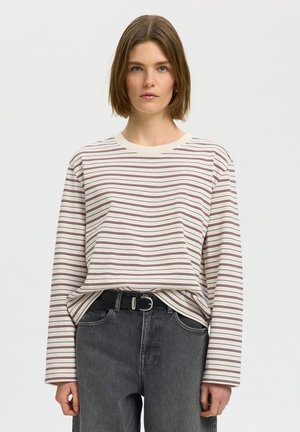 Jeune femme aux cheveux bruns courts portant une chemise blanche à manches longues avec de fines rayures rouges et un jean noir taille haute.