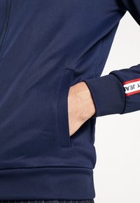 Navy blue zip-up jacket with a smooth texture, featuring side pockets and ribbed cuffs. Red and white stripe accent on the sleeve.