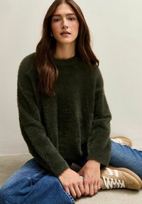 Vrouw met lang bruin haar, zittend in kleermakerszit, gekleed in een donkergroene, pluizige trui, blauwe spijkerbroek en beige sneakers met witte strepen.