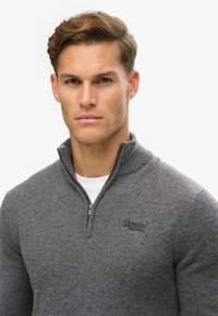 Maglione grigio con zip in morbido tessuto testurizzato, caratterizzato da un alto colletto e un sottile logo sul petto. Indossato sopra una camicia bianca.