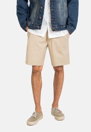 Hombre con pantalones cortos beige, camisa blanca, chaqueta vaquera y zapatillas beige, de pie con las manos en los bolsillos frente a un fondo liso.