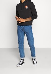 Sudadera negra con estampado gráfico, vaqueros azules y zapatillas altas negras. Los vaqueros están remangados en los puños, mostrando un ajuste relajado.