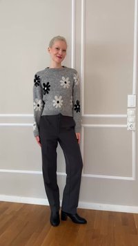 Femeie stând în interior, purtând un pulover gri cu flori negre și albe, pantaloni închiși la culoare și cizme negre cu toc, în fața unui perete bej.