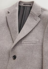 Szary wełniany żakiet z klapami, pojedynczym zamknięciem na czarny guzik i kieszenią na piersi. Wnętrze wyłożone jest wzorzystą podszewką.