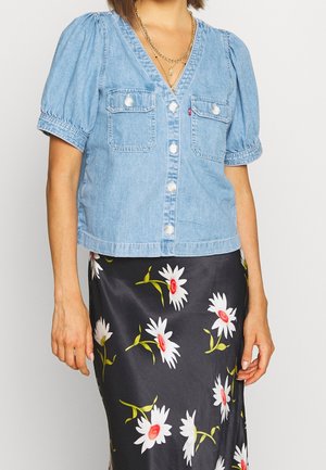 Vrouw draagt een lichtblauw kort mouw denim blouse met parelknopen en een zwarte rok met wit en rood madelief bloemenpatroon.