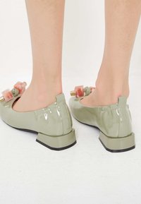 Glanzende, lichtgroene schoenen met een lage blokhak, voorzien van twee decoratieve accenten in het roze en groen aan de voorkant. Zachte textuur.
