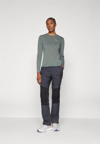 Green long-sleeve top with a small logo, paired with dark blue and black cargo pants featuring zippered pockets and a tapered fit. White athletic shoes.