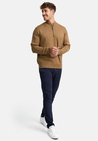 Brauner Strickpullover mit einem halben Reißverschluss, kombiniert mit navyblauen Slim-Fit-Hosen und weißen Sneakers. Einfaches, lässiges Design mit gerippten Bündchen.