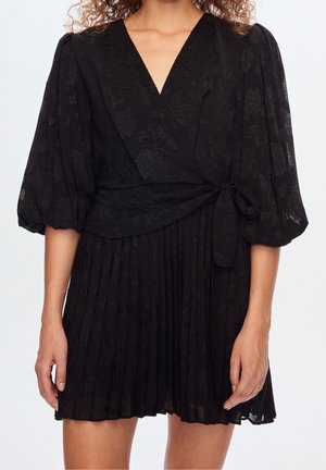 Femme portant une robe portefeuille noire avec manches bouffantes et jupe plissée, ornée de motifs floraux subtils et d’un nœud à la taille.
