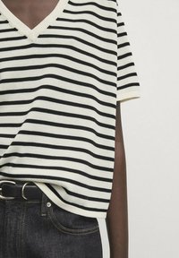 Dzianinowa bluzka z dekoltem w serek i krótkim rękawem w kolorze kremowym z czarnymi poziomymi paskami. Miękki materiał o swobodnym kroju, połączony z ciemnoniebieskimi dżinsami i czarnym paskiem.