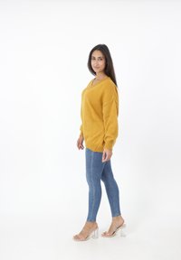 Mosterdgele gebreide sweater met oversized mouwen en V-hals, gecombineerd met blauwe skinny jeans en doorzichtige haksandalen.