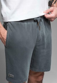 Šedé bavlněné šortky s elastickým pasem na šňůrku, bočními kapsami a malou nášivkou na levém lemu. Měkká textura a pohodlný design.