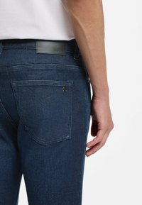 Tummansiniset suorat farkut, joissa on musta nahkainen logopläjäys vyötäröllä ja takataskussa hillitty brändäysyksityiskohta.