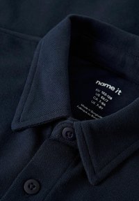 Mornarski polo majica s teksturiranim materijalom, kragnom i dva crna gumba. Oznaka za njegu vidljiva iznutra, s podacima o veličini i podrijetlu.