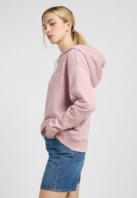 Lichtroze hoodie met een voorzak en een capuchon met trekkoord, gecombineerd met denim shorts. De hoodie heeft subtiele tekst op de voorkant.