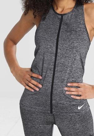 Vrouw draagt een grijze sportoverall zonder mouwen met een zwarte verticale streep en het Nike-logo, handen op de heupen.