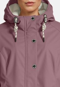 Frau in einer staubigen rosa Kapuzenjacke mit weißem Fleece-Futter und schwarzen Druckknöpfen, ausgestattet mit weißen Kordeln mit schwarzen Spitzen.