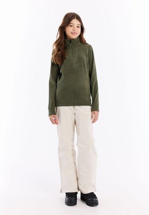 Dunkelgrüner gerippter Fleece-Pullover mit hohem Kragen, kombiniert mit weißen Skihosen und schwarzen Stiefeln. Einfaches Design, minimale Hardware.