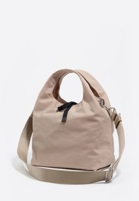Beżowa torba materiałowa z miękkiego materiału z okrągłymi wycięciami na uchwyty, czarną wstążką oraz regulowanym beżowym paskiem na ramię z metalowymi zapięciami.