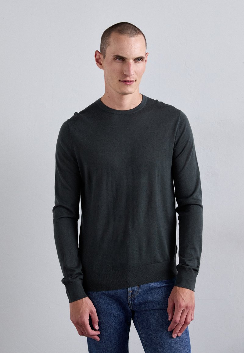 Dunkelgrüner Strickpullover mit Rundhalsausschnitt und langen Ärmeln, mit einem enganliegenden Saum und weicher Textur, kombiniert mit blauen Jeans.