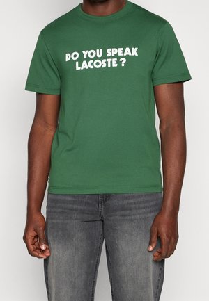 Groen katoenen T-shirt met witte tekst in een vet en speels lettertype: "SPREEK JIJ LACOSTE?" Korte mouwen, ronde hals.