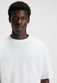 Hvid bomulds T-shirt med rund hals, korte ærmer og et tekstureret "HUGO" logo på venstre bryst. Glat, let stof.