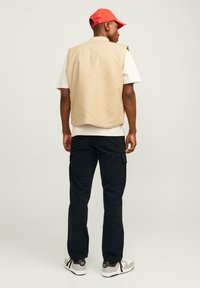 Gilet beige senza maniche su una t-shirt bianca, pantaloni cargo neri e sneakers grigie. Cappellino rosso. Vista posteriore, tessuto liscio, vestibilità comoda.