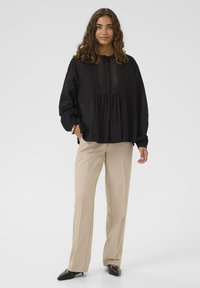 Frau mit lockigem Haar, die eine schwarze Bluse mit langen Ärmeln, beige Hosen und schwarze spitze Schuhe trägt, steht mit einer Hand in der Tasche.