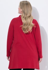 Donna con lunghi capelli biondi e mossi che indossa una tunica rossa a maniche lunghe, in piedi contro un muro bianco, rivolgendosi dalla parte opposta rispetto alla fotocamera.