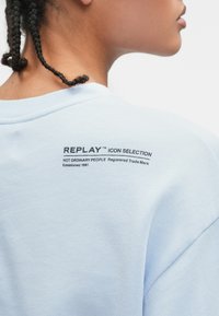 Ανοιχτό μπλε φούτερ με στρογγυλή λαιμόκοψη. Διαθέτει μαύρο εκτυπωμένο κείμενο στην πλάτη, συμπεριλαμβανομένου του ονόματος της μάρκας και του σλόγκαν. Απαλή υφή.