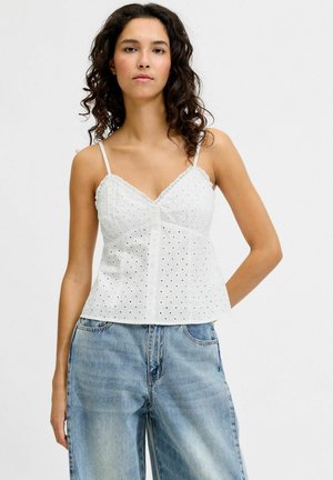 Femme aux cheveux bouclés et foncés portant un haut en dentelle blanche à œillets et un jean taille haute bleu clair, se tenant devant un fond blanc.