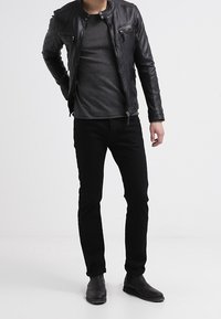 Musta nahkatakki vetoketjullisilla taskuilla, harmaa t-paita, mustat farkut ja mustat nilkkurit. Malli seisoo yksinkertaista valkoista taustaa vasten.