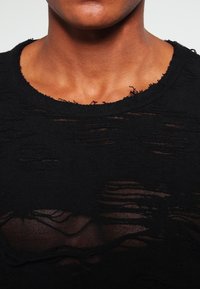 T-shirt noir avec une texture usée, présentant des trous irréguliers et des bords effilochés. Encolure ronde, tissu léger.