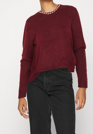 Femme portant un pull bordeaux avec un col orné de perles et un jean noir taille haute, posant devant un fond uni.