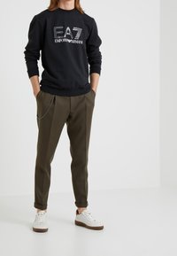 Muž v černé mikině Emporio Armani, olivově zelených kalhotách s řetízkovým detailem a bílých teniskách na jednotném bílém pozad�í.